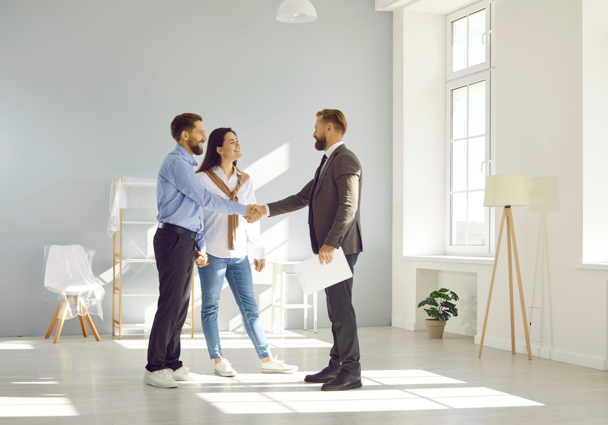 Young married couple make deal, buy new house and exchange handshake with relator. Happy man and woman standing in modern light spacious light living room and shaking hands with real estate agent