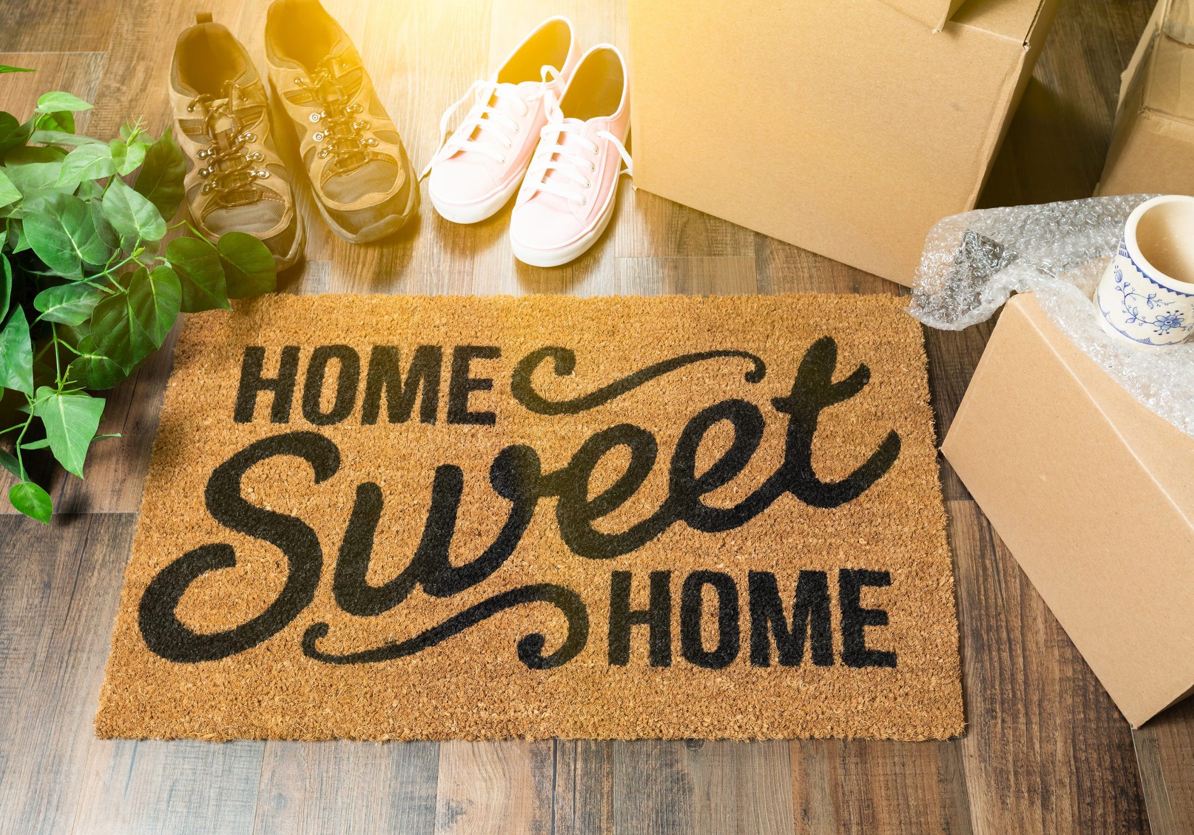 Home Sweet Home Welcome Mat, Moving Boxes, Women and Male Shoes and Plant on Hard Wood Floors.